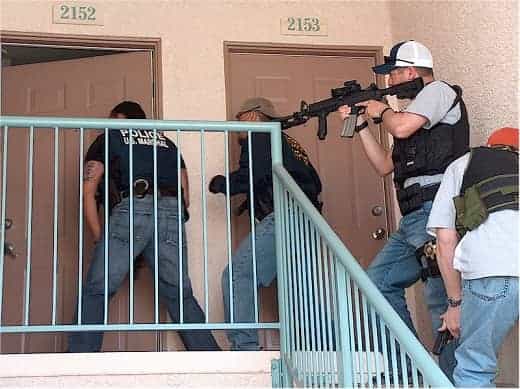 U.S. Marshals, one with a attack rifle, on a stairway approaching a closed door.